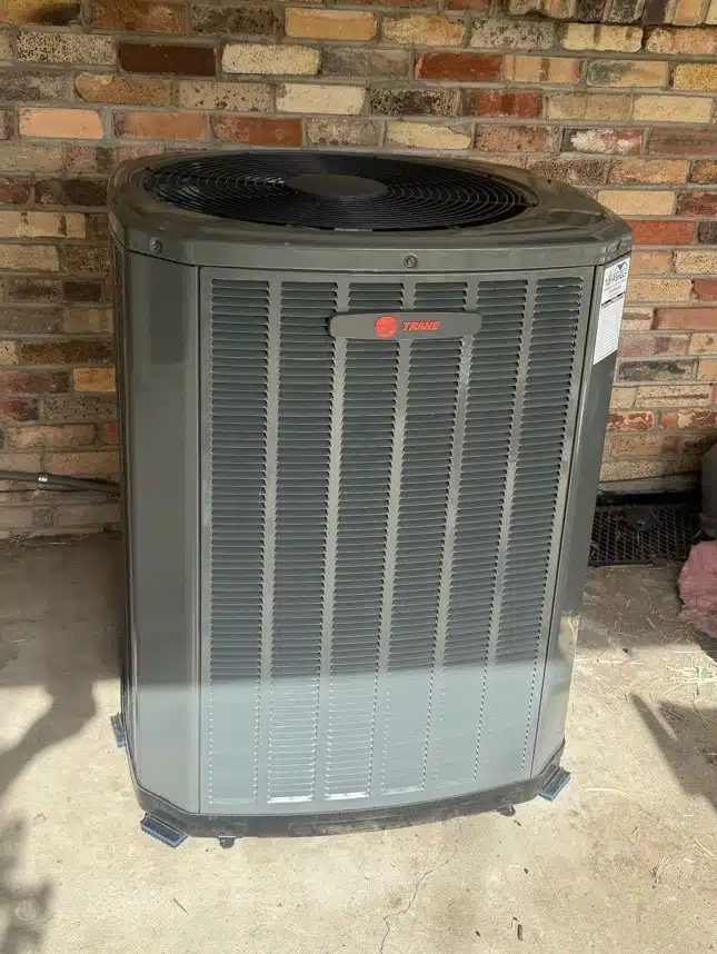 Brand-new Trane AC condenser sitting on adjustable feet against a brick exterior, installed in Denver, CO
